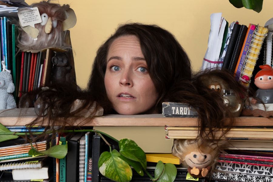 Zoë Coombs Marr’s head is floating in a bookshelf. Her long dark hair is draped over the books and trinkets that cover the shelf.  