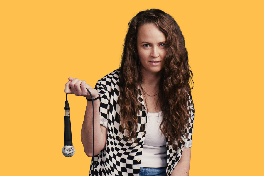 Image is of Zoe Coombs Marr sitting on a stool and dangling a microphone from her left hand. She is wearing a white shirt, blue jeans and a black and white checked top. The background is yellow.