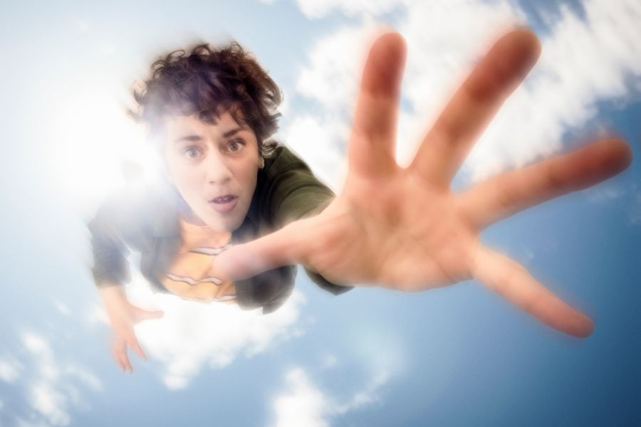 Image is of Jude Perl. She is a Caucasian woman who has short brown, curly hair and brown eyes. She is depicted mid-air, extending her hand toward the camera lens. She is wearing a green jacket over a yellow and white striped t-shirt. She is in front of a bright sky with scattered clouds and sunlight.