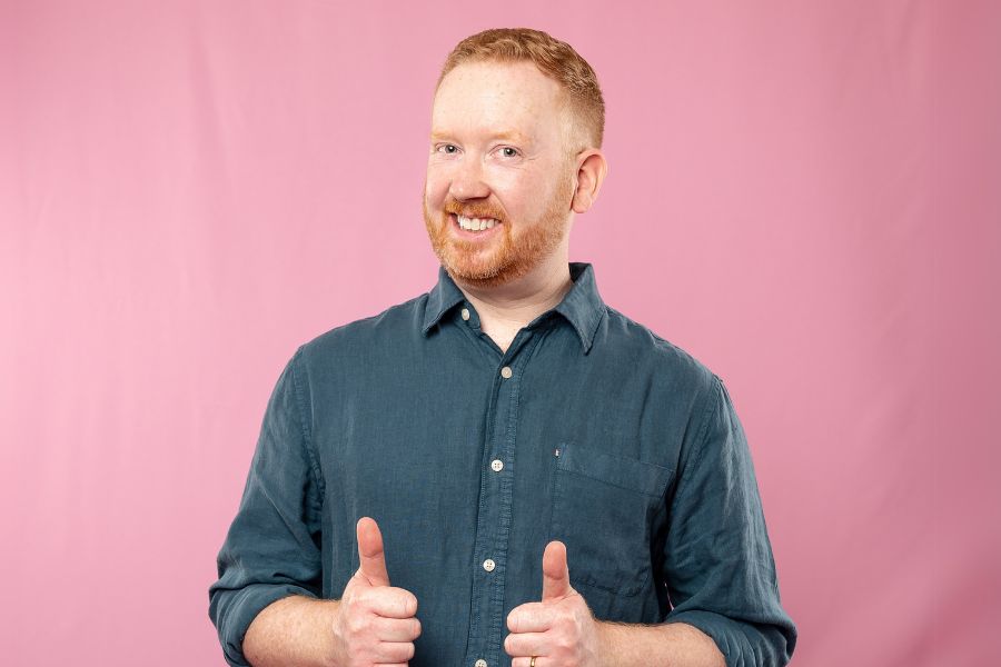 Luke McGregor has short red hair and a beard. He is standing in front of a pink background and is wearing a denim button up. He is smiling with his teeth, has his thumbs up and is looking into the camera.