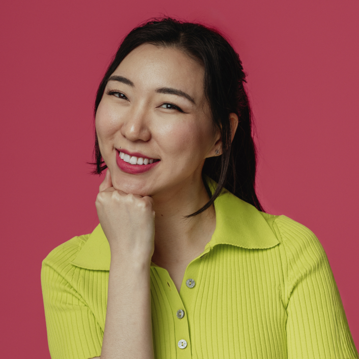 Jenny Tian smiles brightly to the camera with her right hand in a fist and her chin resting on her knuckles, her head gently tiled to her left. Her black, long straight hair is brushed back with two bangs dropping past each ear. She has red lipstick on and small silver ear studs in the shape of a simple flower. She wears a retro lime green nylon collared shirt with vertical textured lines and three buttons, the top one undone. The background is a light maroon.