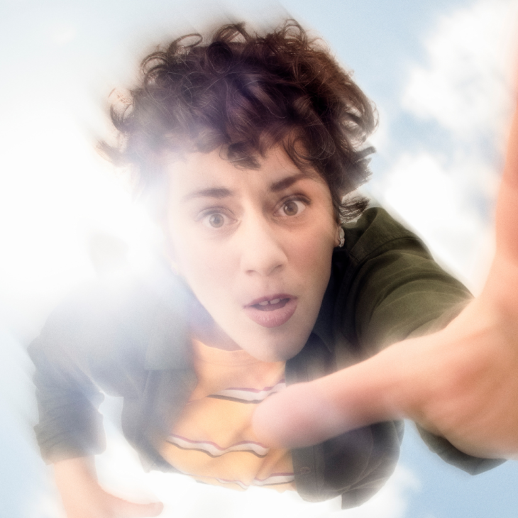 Image is of Jude Perl. She is a Caucasian woman who has short brown, curly hair and brown eyes. She is depicted mid-air, extending her hand toward the camera lens. She is wearing a green jacket over a yellow and white striped t-shirt. She is in front of a bright sky with scattered clouds and sunlight.