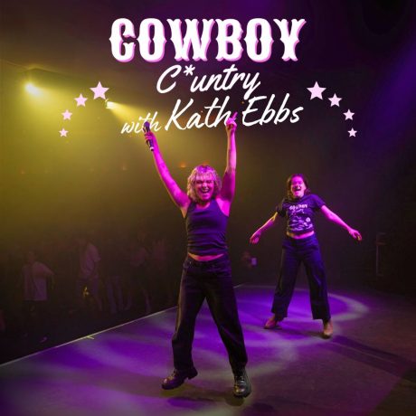 Kath ebbs is on a stage. Pink spotlight on them. They jump with a microphone in hand. People line dance in the background.