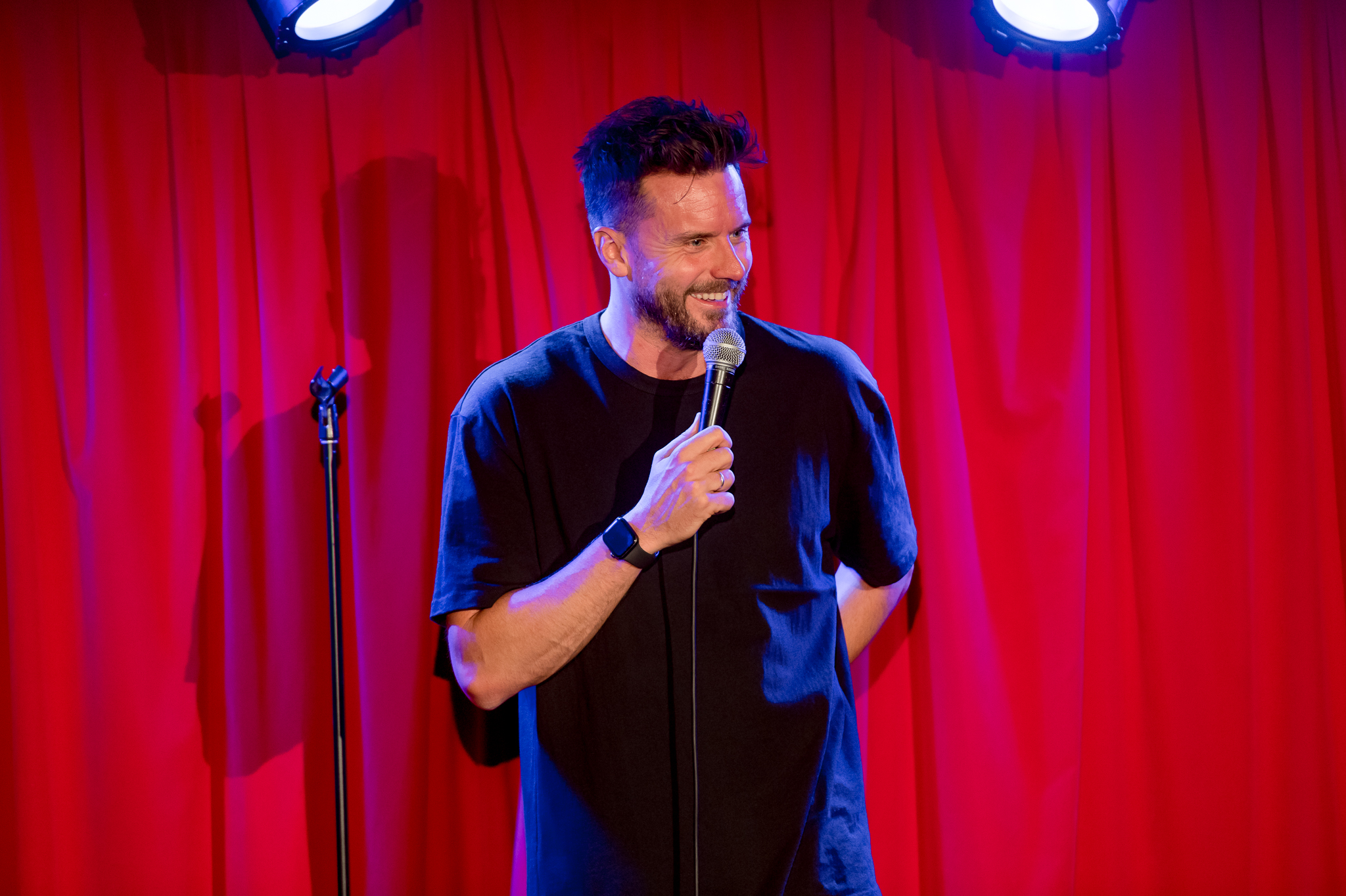 Dave Thornton, a comedian with short dark hair and a beard, is performing on stage in front of a red curtain. He is wearing a black t-shirt and a smartwatch on his left wrist. He is holding a microphone in his right hand and smiling while looking towards the audience. Stage lighting casts a warm glow on him, with blue highlights from the overhead lights.