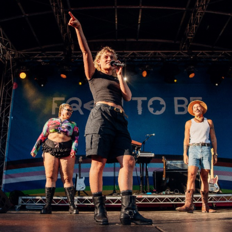 Kath Ebbs stands front centre of stage with their right arm up, the index finger pointing out to the crowd. They wear a black knee length skirt and a black tank top. behind them are a woman and a man smiling, standing wither side of Kath. The background is a stage, with band set-up on a slightly raised platform.