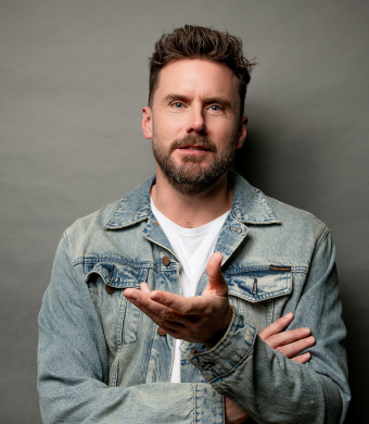 Dave Thornton stands against a neutral gray background. He has short brown hair, a well-groomed beard, and a relaxed posture. He is wearing a light blue denim jacket over a white t-shirt. His left arm is crossed over his chest, while his right hand is extended outward in a gesturing motion.