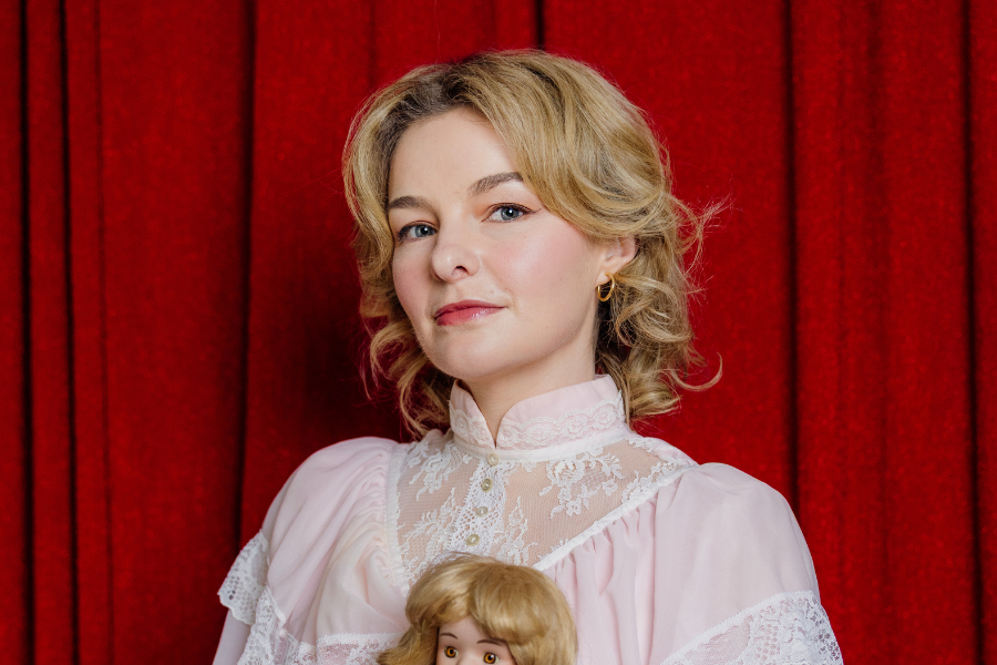 Image is of Abby Howells standing in a pink lacy dress. She has blonde short wavy hair and it holding a doll that looks similar to her. She is standing in front of a red curtain.