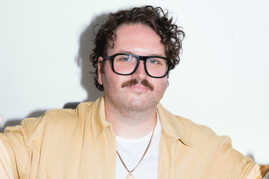 Alexei Toliopoulos has short brown curly hair and a moustache. He is wearing a yellow un-buttoned shirt, white t-shirt and a gold necklace. The background is white.