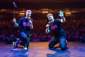 Image is of Wil Anderson and Tom Ballard kneeling on a stage in front of a audience. Both are wearing ASRC Benefit t-shirts.