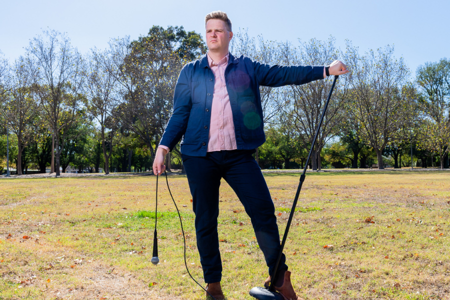 Image is of Tom Ballard standing in a field. He is holding a mic stand in one hand and dangling a microphone in the other. He is wearing black jeans, a pink button shirt and a navy blue jacket.