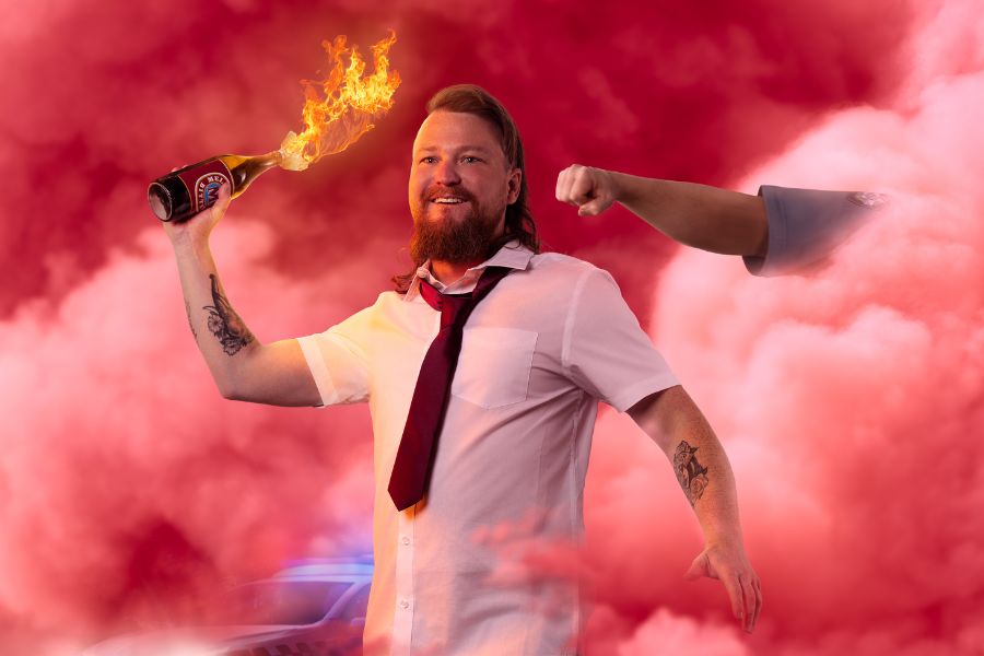 Brett Blake is waring a black shirt and jacket. He has a brown beard and a mullet. The background is grey.