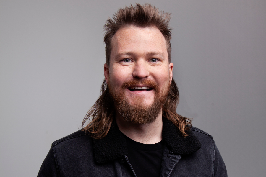 Brett Blake is waring a black shirt and jacket. He has a brown beard and a mullet. The background is grey.