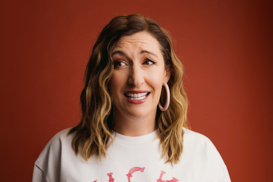 Celia Pacquola has medium length brown wavy hair. She is wearing a white sweatshirt and large pink hoop earrings. She has a worried look on her face and she is looking out of frame. The background is burnt orange.