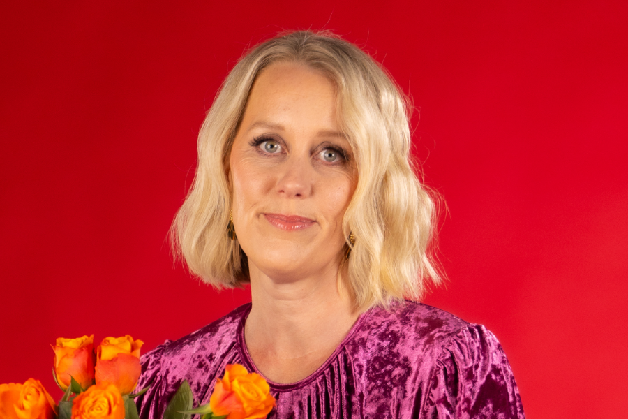 Claire Hooper has short blonde wavy hair. She is wearing a purple patterned dress and is looking into the camera lens and smiling. Claire is holding a bunch of orange roses and is sitting in front of a red background.