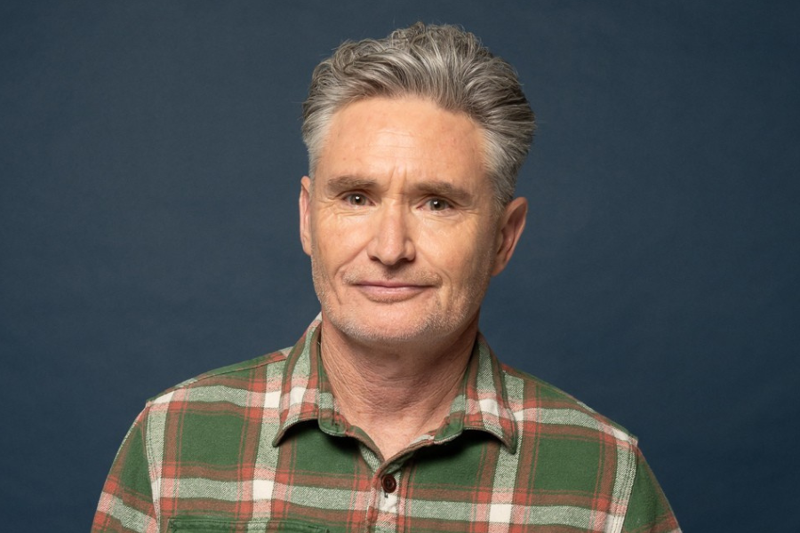 Dave Hughes has short grey hair and is wearing a green and orange checkered shirt. He is standing in front of a blue backdrop and smiling.