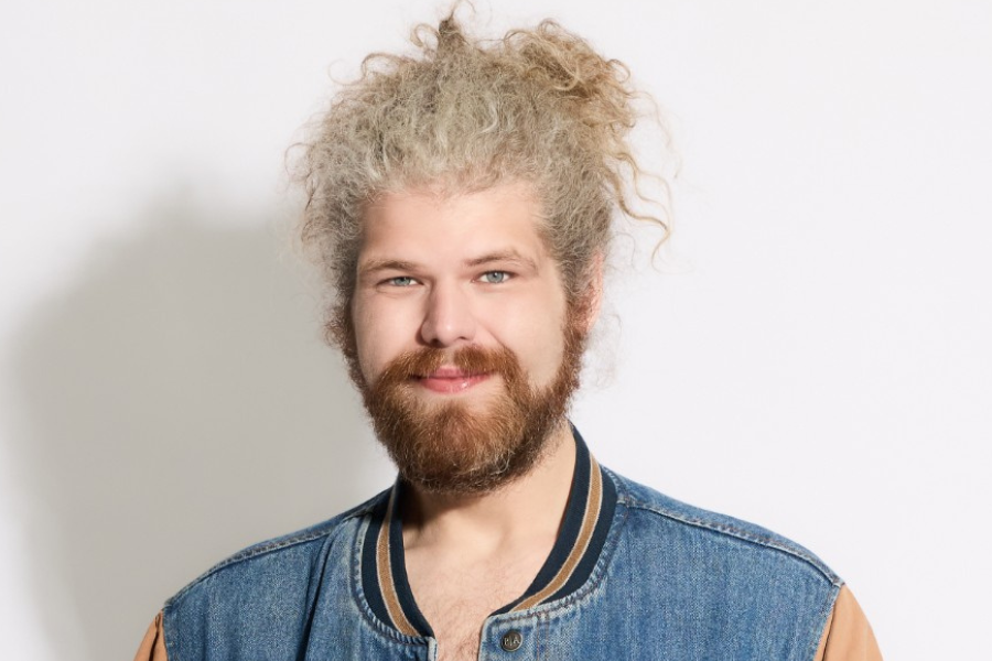 Image is of Dave Woodhead. He has long curly light coloured hair which has been pulled into a bun. He has an orange beard and is wearing a denim jacket with a blue and gold trim. He is standing in front of a red background and smiling.