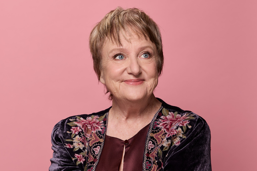 Denise has short blonde hair. She is wearing a silky and velvet textured top that is green, purple, brown, yellow and pink. There is a floral pattern on her shoulders. She is smiling out of the frame.