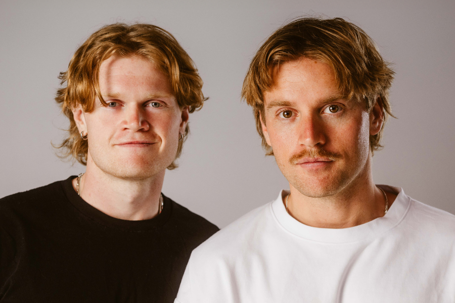 Image is of The Fairbairns. Jaxon is wearing a black tshirt and Lachlan is wearing a white tshirt. Both have blonde wavy hair. The background is grey.