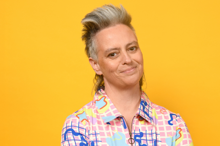 Image is of Geraldine Hickey standing in front of a yellow background and smiling. She has short grey hair and is wearing a colourful buttoned up shirt.