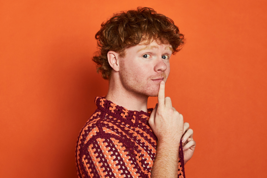 Image is of Wil Gibb. He is wearing a red and orange crocheted top and is posing toward the camera. Wil has red hair and the background is orange.