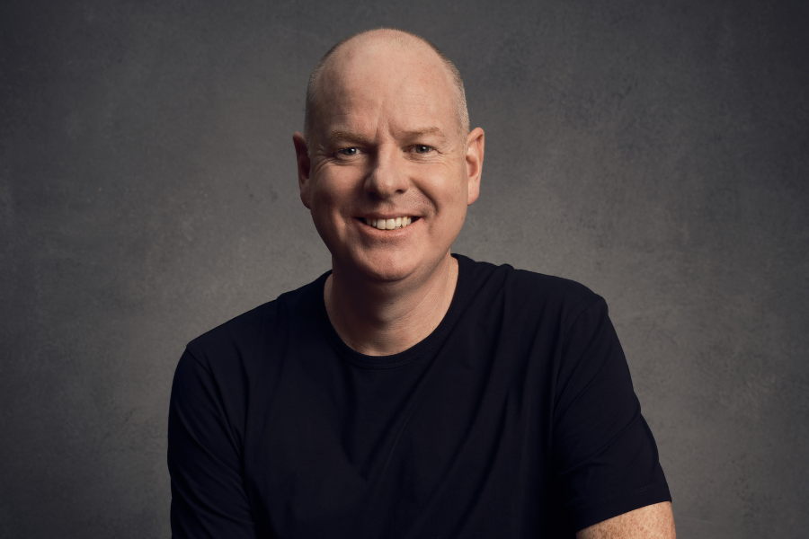 Tom Gleeson sits in front of a grey backdrop. He's wearing a black tshirt and smiling.