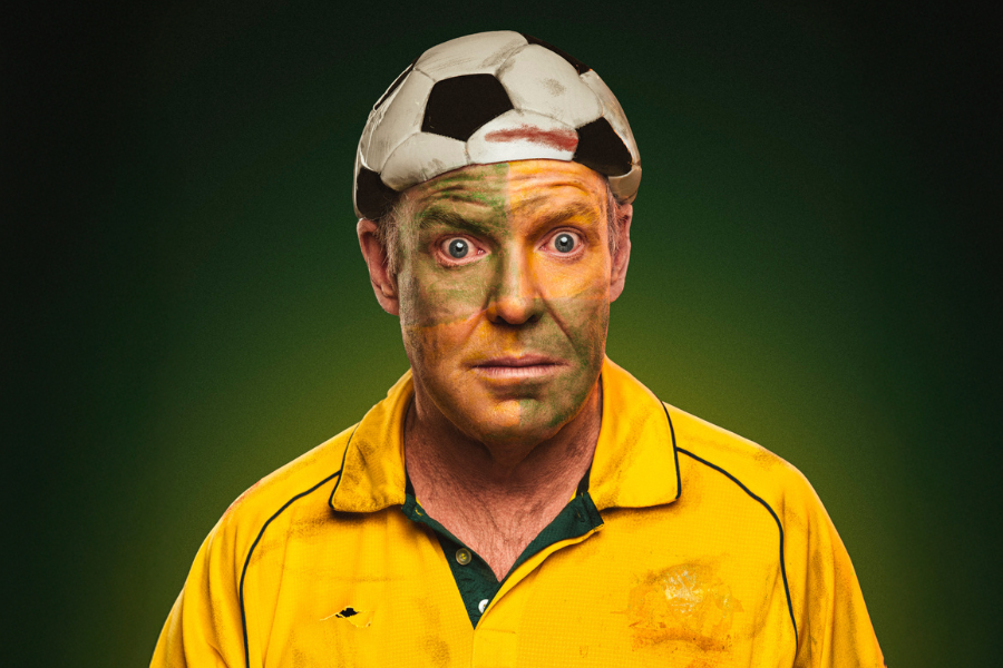 Image is of Pete Helliar. He is dressed in the Australian soccer jersey and has a deflated soccer ball on his head like a hat. He is wearing faded green and yellow face paint and looks disheveled. The background is green.
