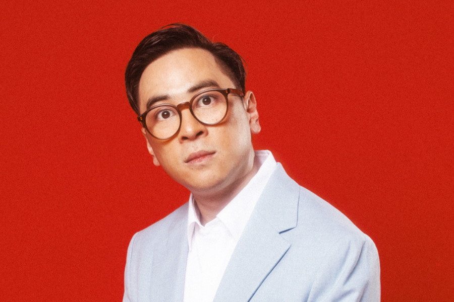 Image is of Michael Hing wearing a grey suit, white shirt and brown reading glasses. He is leaning and looking into the camera. The background is red.