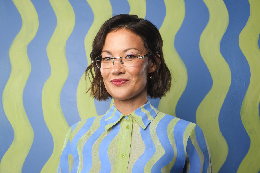 Image is of Lizzy Hoo. She is wearing a green and blue Squiggly shirt. The background is the same pattern as the wallpaper and Lizzy is camouflaging into it. She has short brown hair and glasses.