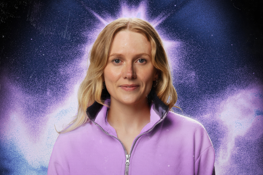 Image is of Jordan Barr who has long wavy blonde hair and is wearing a purple zip up jacket. The background is a purple starry galaxy.