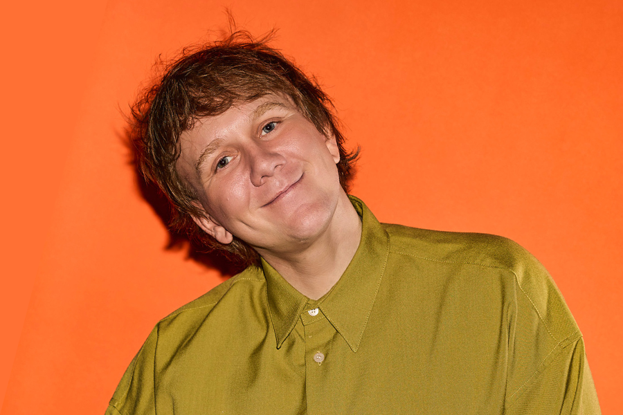 Image is of Josh Thomas wearing a green buttoned up shirt. He has blonde hair and blue eyes. He is leaning sideways and smiling into the camera. He is standing in front of an orange background.