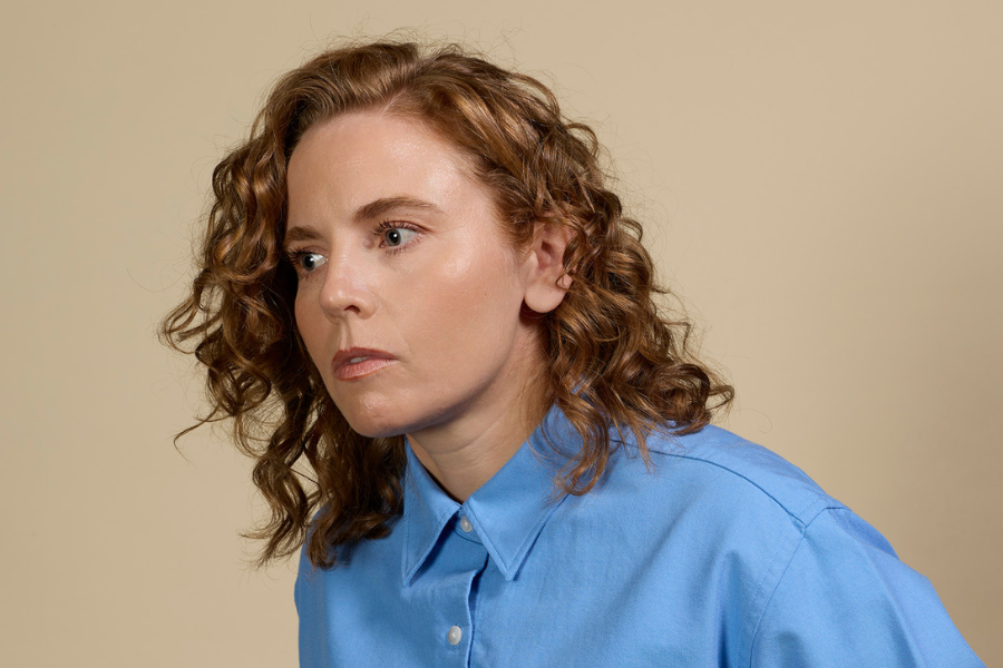 Bronwyn Kuss is wearing a blue button up shirt. She has red curly hair. Her body and face is turning toward the side and she is looking off camera. The background is beige.