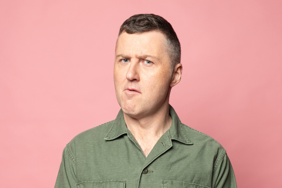 Image is of Lloyd Langford wearing a green button up shirt. He is standing in front of a pink background and has a confused expression on his face.