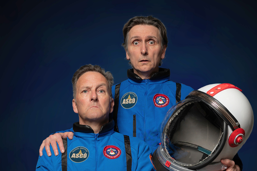 Image is Lano (left) and Woodley (Right). They are both wearing blue and red astronaut uniforms and looking into the camera.