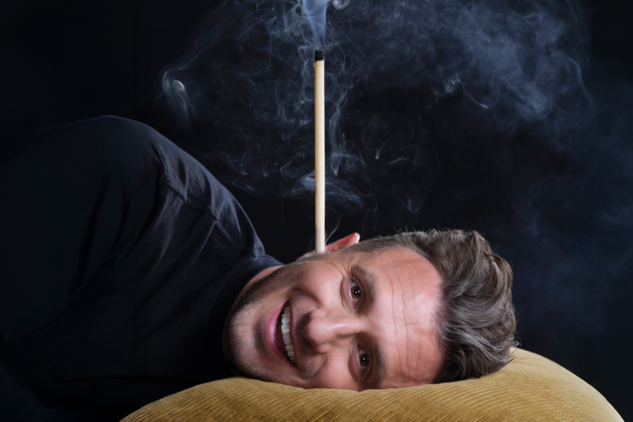 Image is of Tommy Little. Tommy is lying on a mustard coloured corduroy pillow and has an incense coming out of his ear. There is smoke surrounding the image. He is smiling.