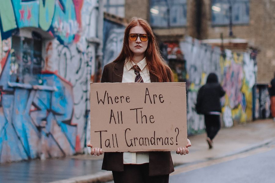 Lou Wall has long orange hair that has been pulled back into a ponytail. They are wearing a green Adidas jacket and a white Adidas top. The background is purple.