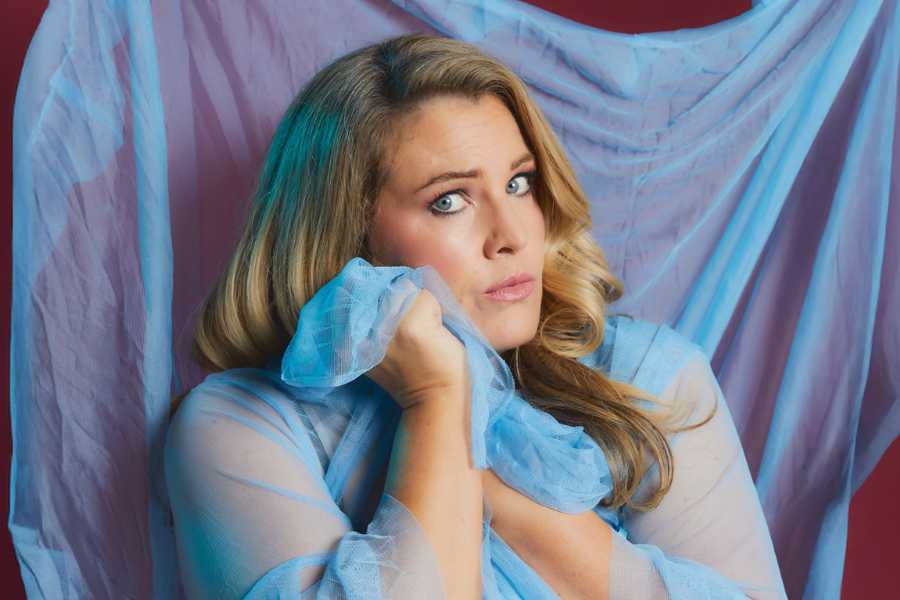 Image is of Nikki Britton. Nikki has long blonde hair and blue eyes. Blue fabric is draped over Nikki and the frame of the image. She is cradling a bit of the fabric and looking into the camera.