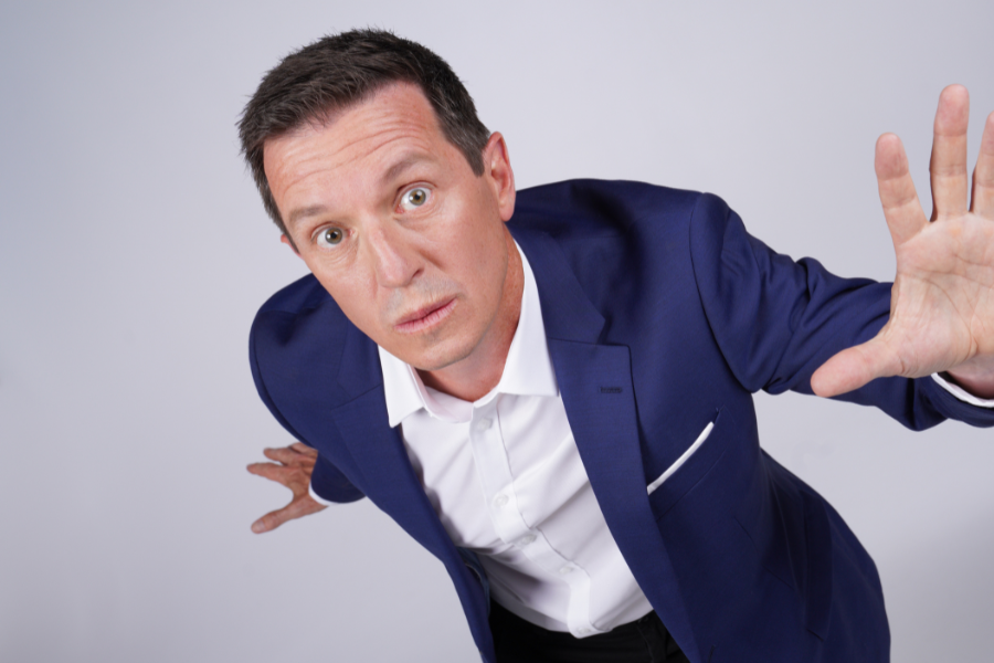 Image is of Rove McManus wearing a blue suit and white shirt. The camera is above Rove and he is reaching up his right hand toward the lens.