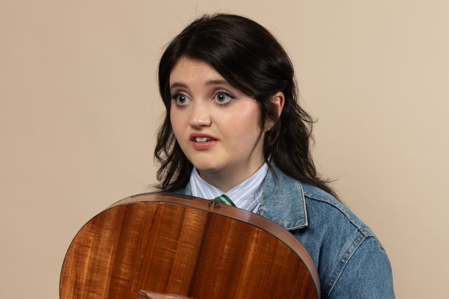Image is of Sez. Sez has long brown hair and big blue eyes. She is wearing a blue and white striped shirt and a denim jacket. She is holding an acoustic guitar and looking into the distance.