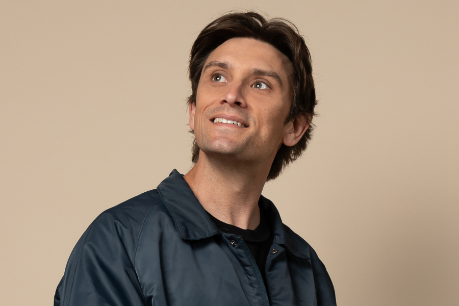 Image is of Sam Taunton. Sam has short brown hair and is earring a navy blue rain jacket. He is looking up to the top left and smiling. The background is light brown.