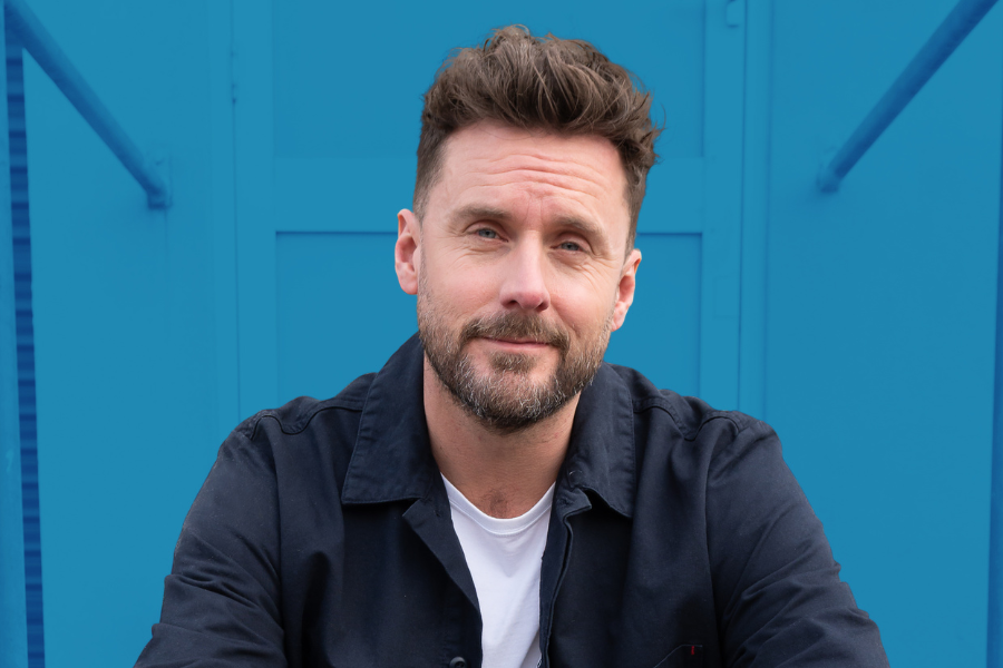 Dave Thornton has short brown hair and a beard. He is wearing a white under shirt and a navy blue buttoned up top. He is sitting in front of a door. The background has been edited to have a blue filter.