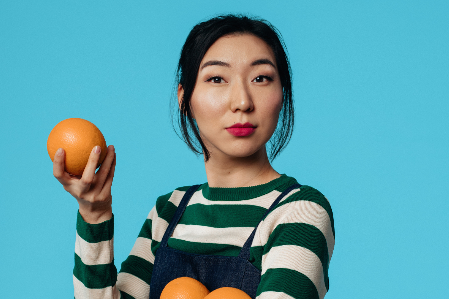 Image is of Jenny Tian wearing a long sleeve green and white striped shirt and navy blue overalls. She has long black hair and red lipstick. Jenny is holding three oranges and looking into the camera.