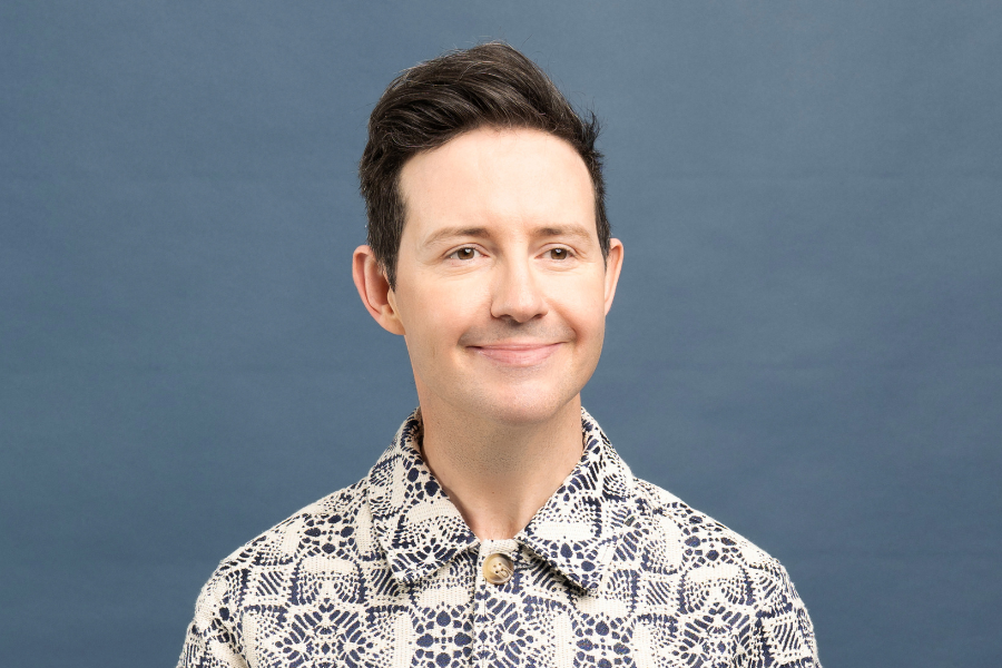 Image is of Nath Valvo wearing a white and blue patterned shirt. He has short brown hair and is smiling toward the side of the camera. The background is a grey blue.