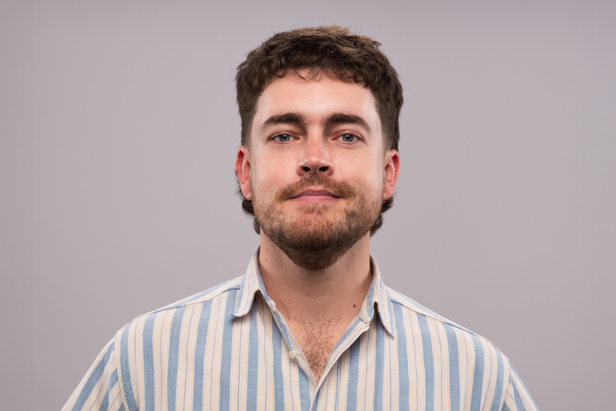 Image is of Nick White wearing a stripy blue and white shirt. He has short brown hair, a beard and blue eyes. He is looking into the camera and smiling without teeth.