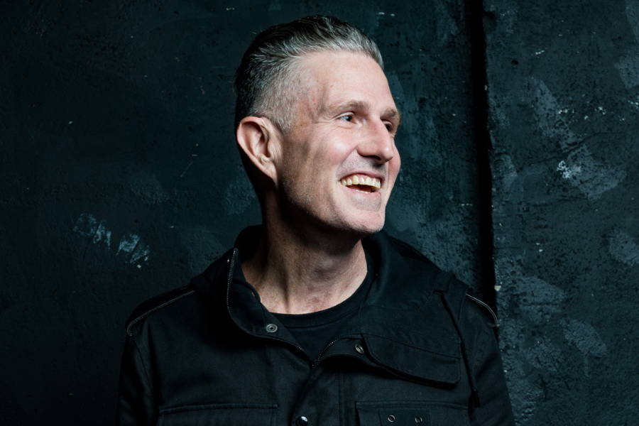 Image is of Wil Anderson. Wil has short grey hair and is wearing a dark jacket. He is smiling and looking to his left.