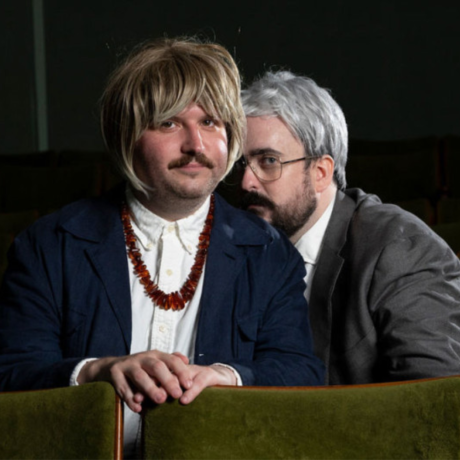 Image is of Zach Ruane and Alexei Toliopoulos. They are seated in a dimly lit theater with green upholstered seats. Alexei is in the foreground. He has a mustache, wears a blonde wig, a dark blue blazer over a white shirt, and a chunky amber-colored necklace. His hands are clasped together on the seat in front of him, and he has a slight smile. Zach, positioned slightly behind Alexei, has a beard, wears a silver-gray wig, glasses, a gray suit, and a white shirt. He leans in close to Alexei, looking intently at him with a serious expression.