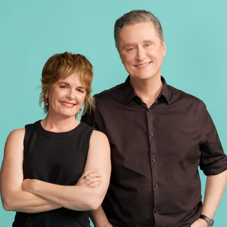 Sarah Kanowski and Richard Fidler are standing together in front of a turquoise background. She has short light brown hair, is wearing a sleeveless black top, is smiling and has her arms crossed. He has short gray hair, is wearing a dark button-up shirt and has one arm behind her.