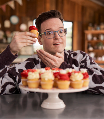 Image is of Tom Walker. He is wearing glasses and a patterned sweater sits at a table in a kitchen. He is holding up a mini cupcake with red frosting and smiling, gesturing to it with his other hand. In front of him is a cake stand filled with similar mini cupcakes decorated with red and white frosting. The background features wooden walls, shelves with jars, and hanging bunting.