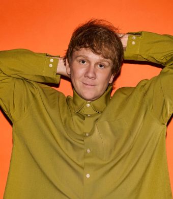 Josh Thomas wears a green shirt and poses on an orange background