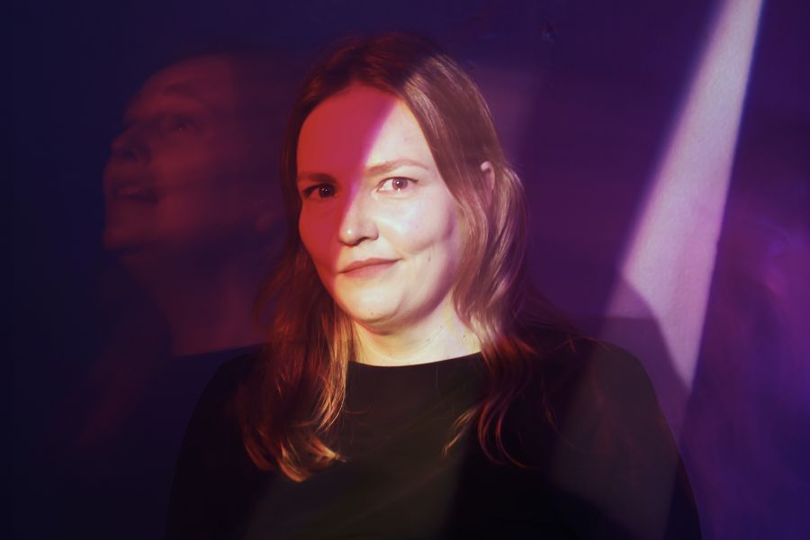 Image is of Geraldine Hickey standing in front of a yellow background and smiling. She has short grey hair and is wearing a colourful buttoned up shirt.