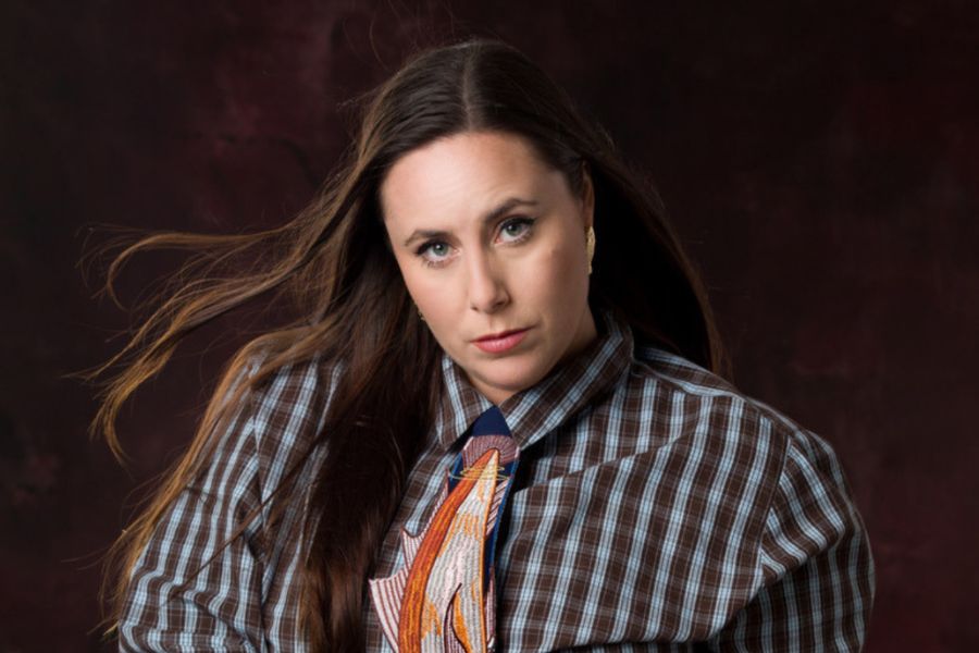 Image is of Geraldine Hickey standing in front of a yellow background and smiling. She has short grey hair and is wearing a colourful buttoned up shirt.
